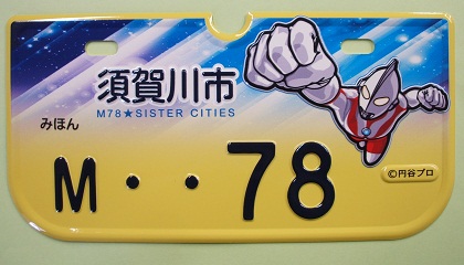 青と白の宇宙のような背景に「須賀川市」と書かれた文字と、拳を突き出し飛んでいるウルトラマンのイラストが描かれた、「M・・78」の黄色のナンバープレートの見本写真