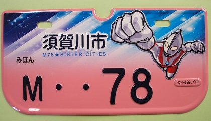 青と白の宇宙のような背景に「須賀川市」と書かれた文字と、拳を突き出し飛んでいるウルトラマンのイラストが描かれた「M・・78」の薄桃色のナンバープレートの見本写真