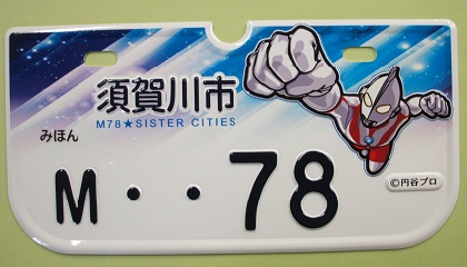 青と白の宇宙のような背景に「須賀川市」と書かれた文字と、拳を突き出し飛んでいるウルトラマンのイラストが描かれた「M・・78」の白色のナンバープレートの見本写真